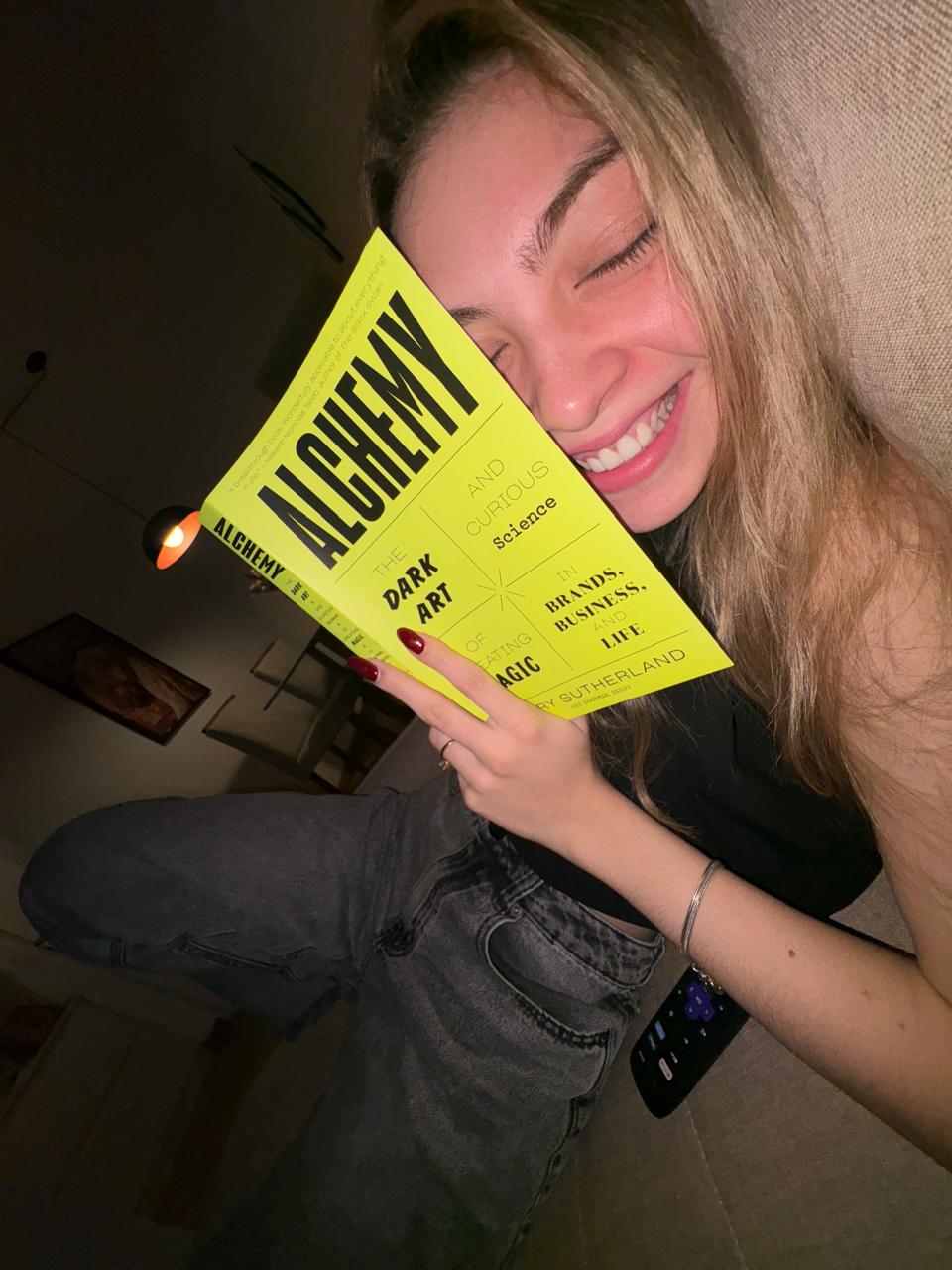 Livia sorrindo, com o livro Alchemy encostado no rosto
